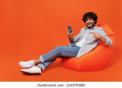 Full size body length young bearded Indian man 20s years old wears blue shirt sit in bag chair hold use pointing index finger on mobile cell phone isolated on plain orange background studio portrait