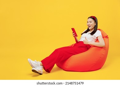 Full Size Body Length Young Girl Woman Of Asian Ethnicity 20s Years Old In Casual Clothes Sit In Bag Chair Hold Use Mobile Cell Phone Show Thumb Up Isolated On Plain Yellow Background Studio Portrait