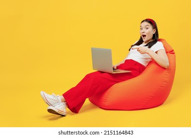 Full Size Body Length Young Girl Woman Of Asian Ethnicity 20s Years Old In Casual Clothes Sit In Bag Chair Hold Point Finger On Laptop Pc Computer Isolated On Plain Yellow Background Studio Portrait