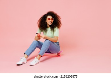 Full size body length young curly latin woman 20s wears casual clothes sunglasses sit on skateboard look aside hold use mobile cell phone isolated on plain pastel light pink background studio portrait - Powered by Shutterstock