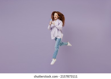 Full Size Body Length Young Redhead Curly Green-eyed Woman 20s Wears White T-shirt Violet Jacket Jump Hold In Hand Use Mobile Cell Phone Isolated On Pastel Purple Color Wall Background Studio Portrait