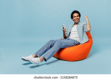 Full size body length young black curly man 20s wears white shirt sit in bag chair hold in hand use mobile cell phone do winner gesture isolated on plain pastel light blue background studio portrait - Shutterstock ID 2158284873
