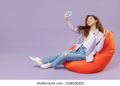 Full size body length young redhead curly woman 20s wears white T-shirt violet jacket sit in bag chair do selfie shot on mobile cell phone isolated pastel purple color wall background studio portrait