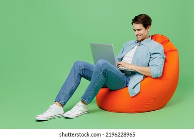 Full size body length young brunet man 20s wears blue shirt sit in bag chair hold use work on laptop pc computer typing browsing chatting send sms isolated on plain green background studio portrait - Powered by Shutterstock