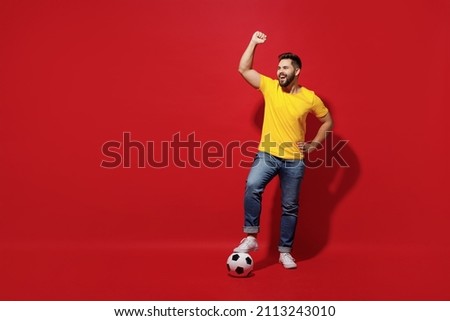 Full size body length young bearded man football fan in yellow t-shirt cheer up support favorite team hold foot on soccer ball do winner gesture isolated on plain dark red background studio portrait