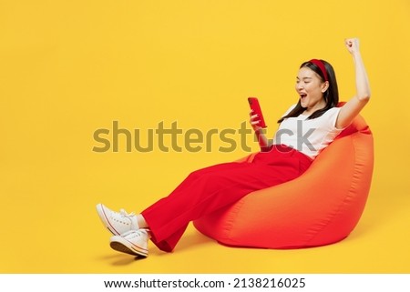 Full size body length young girl woman of Asian ethnicity 20s years old in casual clothes sit in bag chair hold use mobile cell phone do winner gesture isolated plain yellow background studio portrait