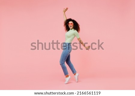Full size body length young curly latin woman 20s wears casual clothes sunglasses stand on toes dance lean back have fun spreading hands isolated on plain pastel light pink background studio portrait