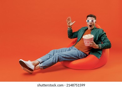 Full size body shoked fun young brunet man 20s wears red t-shirt green jacket sit in bag chairin 3d glasses watch movie film hold bucket of popcorn isolated on plain orange background studio portrait.