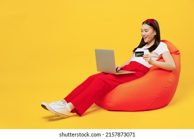 Full Size Body Young Woman Of Asian Ethnicity 20s Years Old Wears Casual Clothes Sit In Bag Chair Use Laptop Pc Computer Hold Credit Bank Card Isolated On Plain Yellow Background Studio Portrait