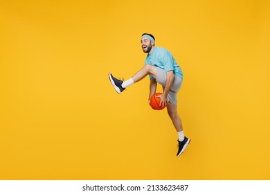 Full Size Caucasian Young Fitness Trainer Instructor Sporty Man Sportsman In Headband Blue T-shirt Hold Ball Play Basketball Game Jump High Isolated On Plain Yellow Background. Workout Sport Concept