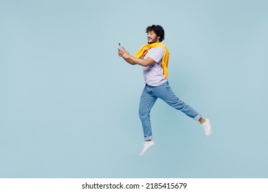 Full Size Excited Young Bearded Indian Man 20s Wears White T-shirt Hold In Hand Use Mobile Cell Phone Typing Browsing Chatting Jumping Isolated On Plain Pastel Light Blue Background Studio Portrait