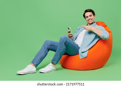 Full size excited young brunet man 20s years old wears blue shirt sit in bag chair hold in hand use mobile cell phone pointing index finger on screen isolated on plain green background studio portrait - Powered by Shutterstock