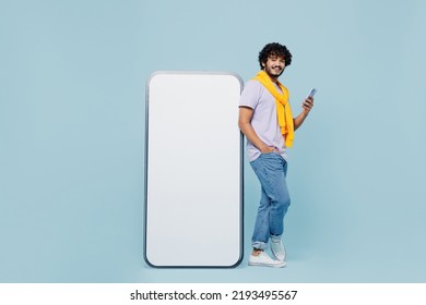 Full size fancy young bearded Indian man 20s wears white t-shirt hold stand near big mobile cell phone with blank screen workspace area isolated on plain pastel light blue background studio portrait - Shutterstock ID 2193495567