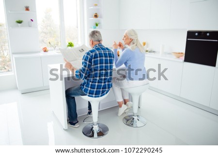 Full size, fullbody rear view portrait of sweet attractive lovely stylish couple sitting at the table in kitchen, man reading magazine, woman having mug of tea, enjoying morning time together