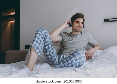 Full Size Fun Happy Smiling Young Man 20s Wear Grey T-shirt Blue Pajamas Headphones Sit In Bed Listen To Music Rest At Home On Weekends Relax Indoors Bedroom Good Mood Night Morning Bedtime Concept