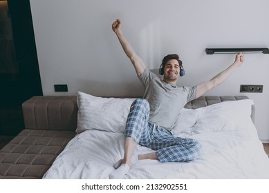 Full Size Fun Happy Young Man Wear Grey T-shirt Blue Pajamas Headphones Sit In Bed Listen To Music Do Stretching Rest At Home On Weekends Relax Indoors Bedroom Good Mood Night Morning Bedtime Concept