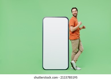 Full Size Fun Young Man 20s Wear Orange T-shirt Stand Point Finger On Mobile Cell Phone With Blank Screen Workspace Area Isolated On Plain Pastel Light Green Color Background People Lifestyle Concept