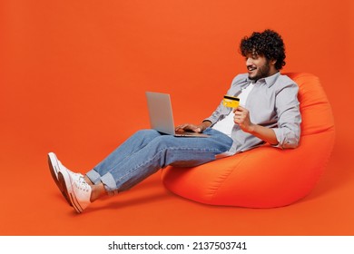 Full Size Happy Young Bearded Indian Man 20s Wears Blue Shirt Sit In Bag Chair Using Laptop Pc Computer Hold Credit Bank Card Doing Online Shopping Isolated On Plain Orange Background Studio Portrait