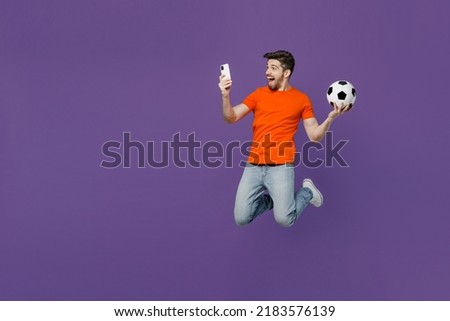 Full size happy young fan man he wears orange t-shirt cheer up support football sport team hold soccer ball use mobile cell phone watch tv live stream jump up isolated on plain dark purple background