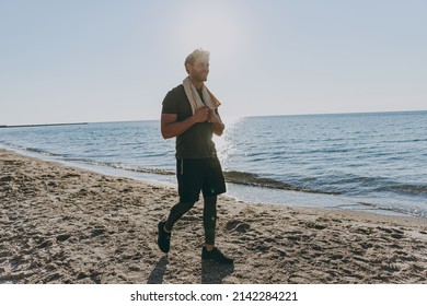 Full size happy young strong athletic toned fit sportsman man in sports clothes towel warm up training walking look aside at sunrise sun dawn over sea sand beach outdoor seaside in summer day morning.