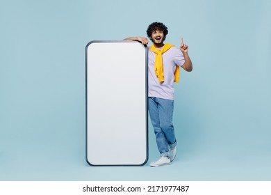 Full size insighted young bearded Indian man 20s wears white t-shirt stand near big mobile cell phone with blank screen workspace area isolated on plain pastel light blue background studio portrait