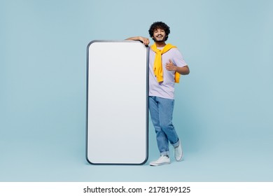 Full size jubilant fun young bearded Indian man 20s wears white t-shirt stand near big mobile cell phone with blank screen workspace area isolated on plain pastel light blue background studio portrait