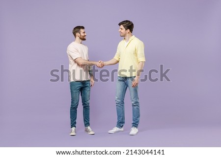 Full size length two young smiling happy men friends together in casual t-shirt meeting together greeting hold hands folded handshake gesture isolated on purple background studio Tattoo translate fun