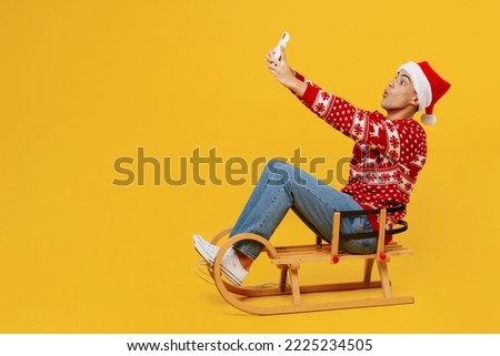 Full size merry young man wear red knitted Christmas sweater Santa hat posing sledding doing selfie shot on mobile cell phone isolated on plain yellow background. Happy New Year 2023 holiday concept