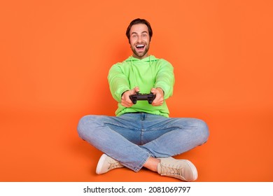 Full size photo of brunet millennial guy play station sit wear green sweatshirt jeans sneakers isolated on orange color background