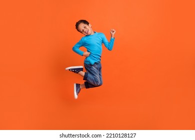 Full size photo of charming small pupil boy jump play raise fists celebrate dressed stylish blue look isolated on orange color background - Shutterstock ID 2210128127