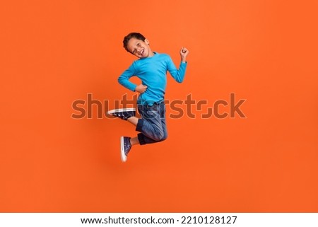 Full size photo of charming small pupil boy jump play raise fists celebrate dressed stylish blue look isolated on orange color background