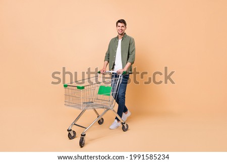 Full size photo of charming young happy man hold pushcart smile shopper isolated on beige color background