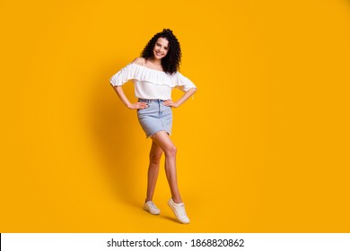 Full Size Photo Of Cheerful Flirty Brunette Woman Standing Hands On Hips Wear White Top Blue Skirt Sneakers Isolated On Yellow Background