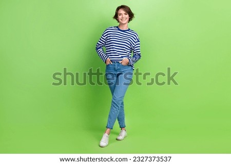 Full size photo of cheerful girl dressed striped sweatshirt standing legs crossed hands in pockets isolated on green color background
