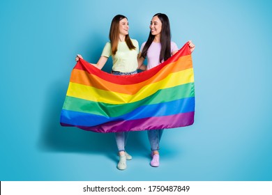 Full Size Photo Of Cheerful Lesbians Couple Lovers Walk Foreign Parade Support Tolerance Same Sex Marriages Carry Gay Rainbow Flag Wear T-shirts Jeans Shoes Isolated Blue Color Background