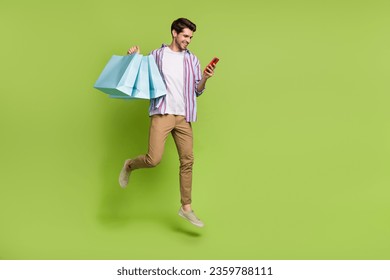 Full size photo of cheerful person flying hold shopping bags look at smartphone order new clothes isolated on green color background