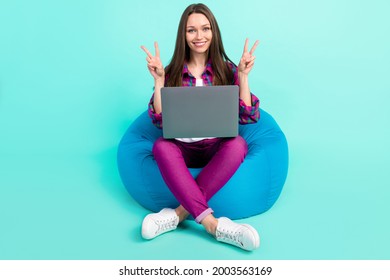 Full size photo of cool young lady sit on bag show v-sign with laptop wear shirt trousers isolated on teal color background