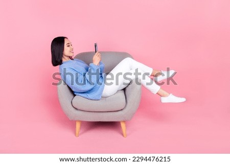 Full size photo of good mood relaxed woman knit pullover trousers look at smartphone sit on arm chair isolated on pink color background