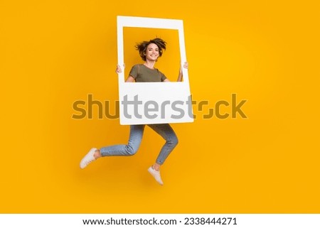 Full size photo of good mood pretty cute girl dressed jeans khaki top hold big photo frame jumping isolated on yellow color background