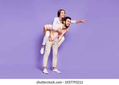 Full Size Photo Of Happy Young Couple Man Piggyback Woman Point Finger Empty Space Isolated On Purple Color Background