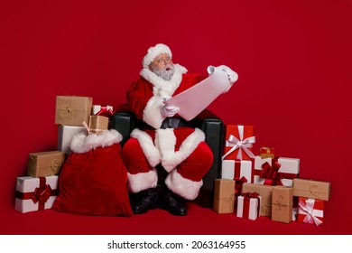 Full size photo of hooray santa sit read letter near presents wear cap eyewear coat trousers shoes isolated on red background - Powered by Shutterstock