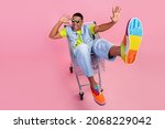 Full size photo of impressed millennial brunet guy sit in trolley wear eyewear t-shirt vest jeans shoes isolated pink background