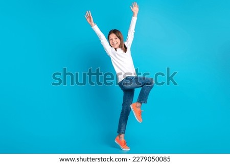 Full size photo of impressed optimistic girl wear white long sleeve denim trousers raising arms up isolated on blue color background