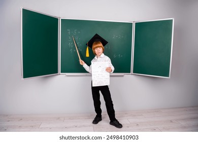 Full size photo of little pupil boy diligent graduation hat teacher hold pointer stick wear trendy uniform classroom blackboard background - Powered by Shutterstock