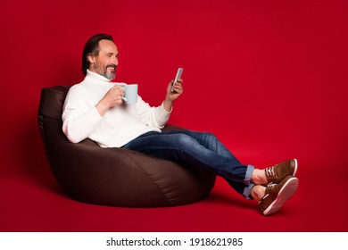 Full size photo of mature happy positive man sit armchair drink tea browsing in internet phone isolated on red color background - Powered by Shutterstock