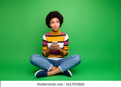 Full size photo minded afro american girl sit legs crossed folded use cell phone blogging think thoughts type social media post wear denim jeans clothes isolated green color background