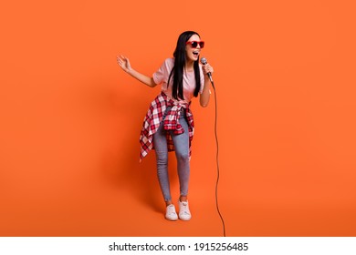 Full size photo of optimistic brunette lady stand sing in mic wear spectacles t-shirt jeans isolated on orange background