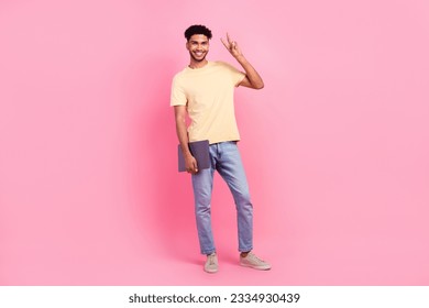 Full size photo of optimistic funny nice man wear yellow t-shirt jeans showing v-sign holding laptop isolated on pink color background