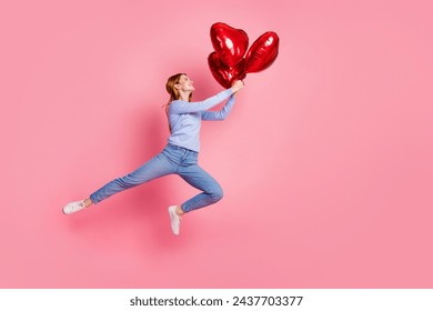 Full size photo of optimistic girl wear blue pullover fly away with red heart helium balloons in arms isolated on pink color background