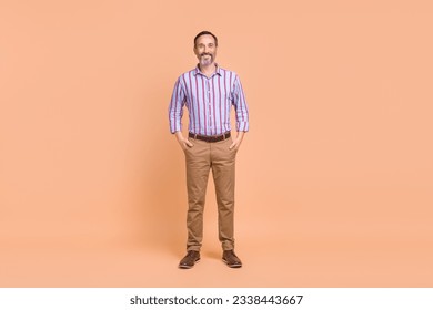 Full size photo of optimistic handsome man wear striped shirt brown pants hold arms palms in pockets isolated on beige color background - Shutterstock ID 2338443667